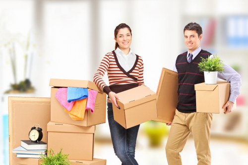 A professional house removal team, consisting of a woman and a man, are indoors in a bright, modern living space with large windows and minimal furnishings. The woman, dressed in a striped jumper and dark jeans, is smiling and holding a medium-sized cardboard box, while the man, wearing a dark sweater with a collared shirt underneath and beige trousers, is also smiling and holding two smaller cardboard boxes, one of which contains a potted green plant. Nearby, several unopened cardboard boxes of various sizes are stacked, some with colorful towels draped over them, and a small black alarm clock is visible on top of one of the boxes. The scene suggests an ongoing moving or packing process typically managed by Bayswater Man with Van, a local removals company serving residential areas in London, with the background featuring a softly blurred living room environment to convey a professional and efficient house removal service.