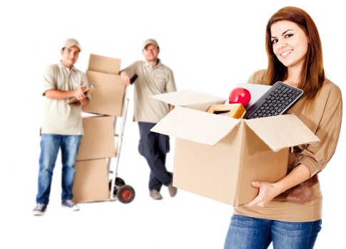 The image features three individuals involved in moving or house removals, set against a plain white background. On the right, a young woman with long brown hair, dressed in a beige top and blue jeans, is smiling while holding an open cardboard box filled with various household items including a red apple, a black remote control, and other small items. Behind her, two men are standing further back, also engaging in packing activities; one is wearing a light beige cap and a matching beige polo shirt, holding a closed cardboard box while the other, dressed similarly, is holding a hand truck with a tall stack of flattened cardboard boxes. The scene represents a professional home removals or relocation service, with a focus on packing and transportation, subtly aligning with the Bayswater Man with Van services provided in the London boroughs, specifically within the postcode area. The lighting indicates a bright, evenly lit environment, emphasizing the organizational process involved in house moving tasks, with all items and individuals clearly visible and positioned in a manner that highlights their roles in the moving process.