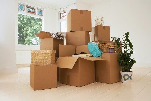 A collection of packed cardboard boxes of various sizes, some open and partially flattened, arranged on a light wooden floor inside a bright, empty room with white walls. The boxes contain household items such as a blue fabric item, a wicker basket, and some smaller wrapped packages. In the background, a large window with white trim and a decorative stained-glass transom allows natural light to illuminate the space, revealing green foliage outside. A potted plant with broad, dark green leaves sits to the right of the boxes in a patterned ceramic pot, while a white radiator is visible beneath the window. The scene appears to depict the process of packing or moving, consistent with house removals services in the Bayswater or central London area, with the company's name, Bayswater Man with Van, implied to be involved in the activity. The setup emphasizes an interior space ready for transition, highlighting household items and the logistics of a move.