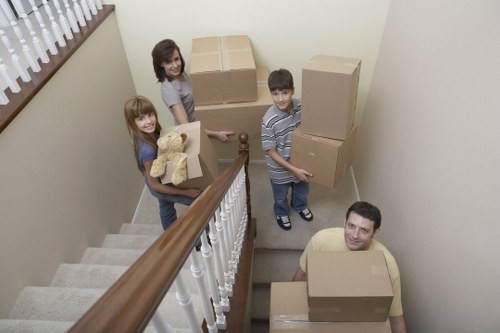 A family of four, including a woman with shoulder-length brown hair, a young girl with long red hair holding a teddy bear and a cardboard box, a young boy with dark hair carrying a large box, and an adult man with short dark hair seated on a staircase, all inside a residential property in Bayswater. The staircase features a wooden handrail and white spindles, with beige carpeted stairs leading upwards. The interior walls are painted in a light cream or off-white tone, and natural light filters in from a window with a lace curtain on the left side. The group is surrounded by packed cardboard boxes of various sizes, some stacked on the stairs and others being carried, indicating an active house move. The scene captures the process of house removals, with household items such as boxes and a teddy bear, and the setting subtly reflects an area within central London near Bayswater with typical residential features. Bayswater Man with Van offers professional removals services, as illustrated by the family preparing for their move.